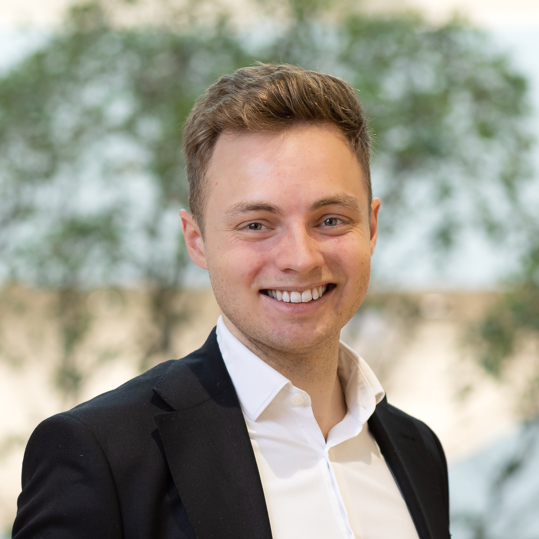 Smiling person in a black blazer and white shirt against a blurred background of greenery.