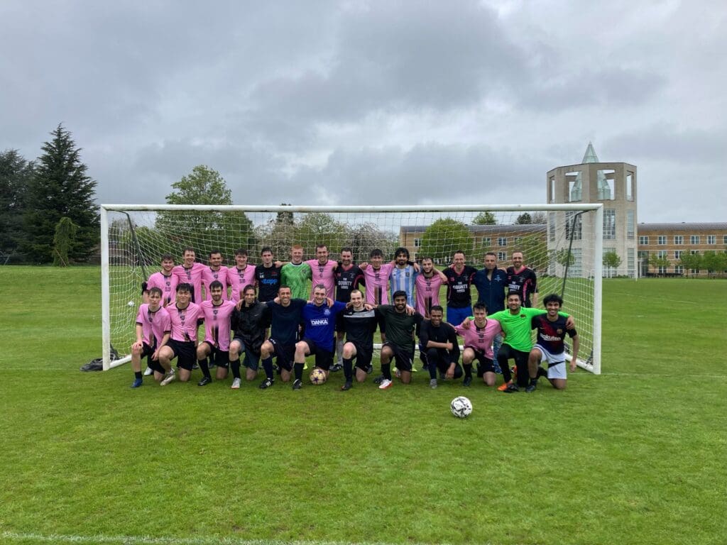 A group of soccer players in pink and black uniforms pose together in front of a goal on a grassy field. Trees and a tall, beige building with arches are visible in the background. Its an overcast day.