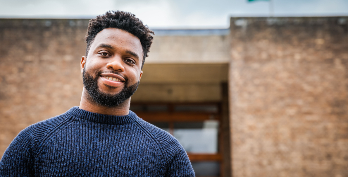 A man with short curly hair and a beard smiles at the camera, wearing a dark blue sweater. They are standing in front of a brick building with a wooden door. The sky is slightly overcast.