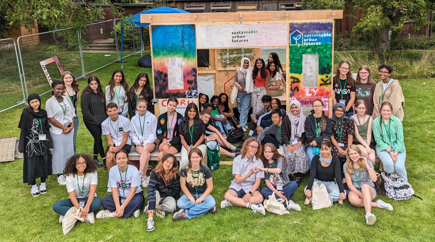 A diverse group of young people posing in front of a wooden structure decorated with colorful posters on a grassy area. The posters read sustainable urban futures. They are smiling and wearing casual clothing.