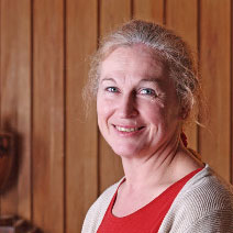 Woman with gray hair tied back, smiling softly. She wears a red top and beige cardigan, standing in front of a wood-paneled wall.