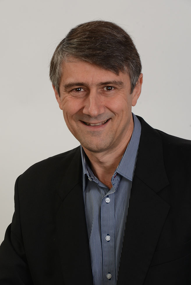 Man with short gray hair, wearing a dark blazer and a blue-striped shirt, smiles while looking at the camera. White background.