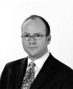 Black and white portrait of a man in a suit and tie, wearing glasses. The person has short hair and is looking directly at the camera with a neutral expression.