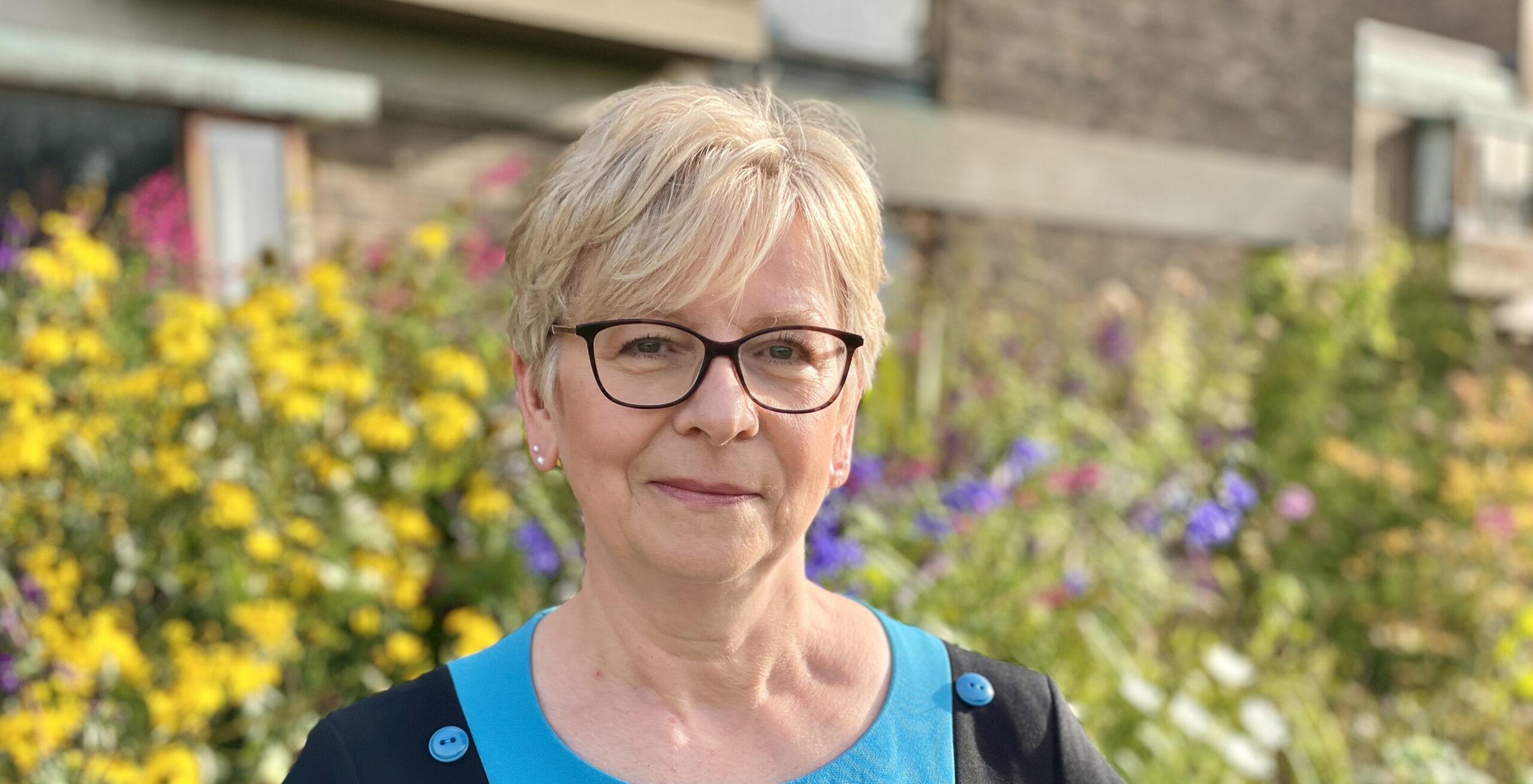 A woman with short blond hair and glasses stands outside, wearing a blue top with black accents. They are in front of a blurred background of colorful flowers and greenery.