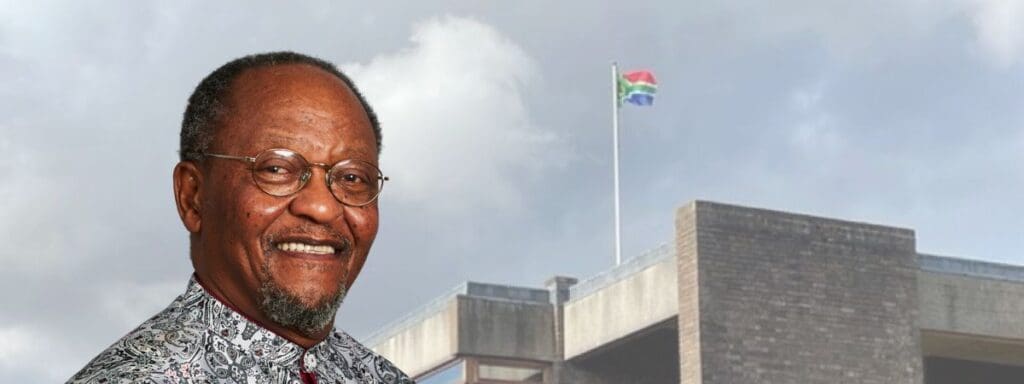 A smiling person wearing glasses and a patterned shirt in the foreground. In the background, a South African flag is flying atop a building against a cloudy sky.