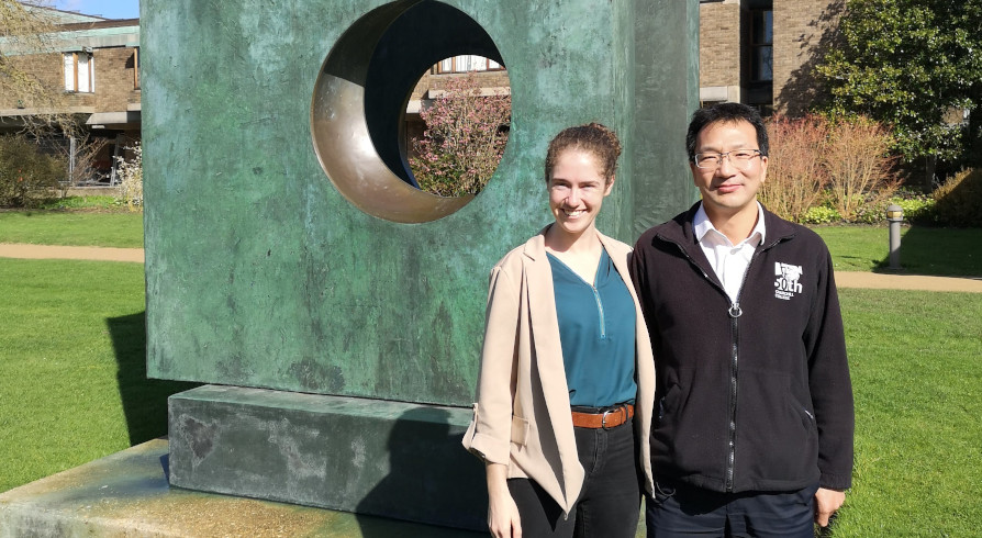 Two people stand in front of a large, abstract bronze sculpture with a circular opening. They are smiling and dressed in casual attire. Green grass and a building are visible in the background.