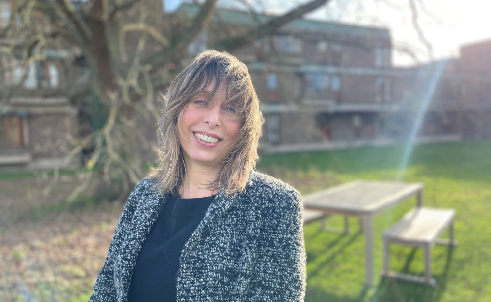 A woman with shoulder-length hair, wearing a patterned gray jacket, smiles while standing outdoors. The sun shines brightly, casting light on their hair. A large tree and building are in the blurred background with picnic tables nearby.