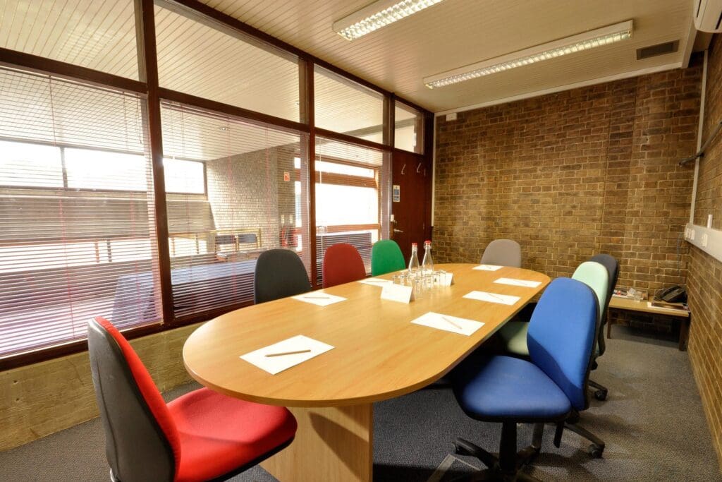 A conference room with a wooden table surrounded by six colorful chairs. The table has paper, pens, and water bottles on it. The room has a brick wall and glass partitions with blinds, allowing natural light to filter through.