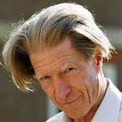 A man with a thoughtful expression and swept-back gray hair is looking slightly downward. They are wearing a white shirt, and the background is blurred.