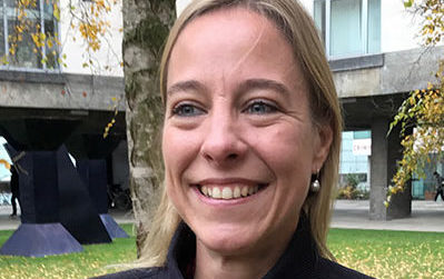 A woman with shoulder-length blonde hair smiling outdoors. She is wearing a dark jacket and pearl earrings. Trees and a modern building are in the background.