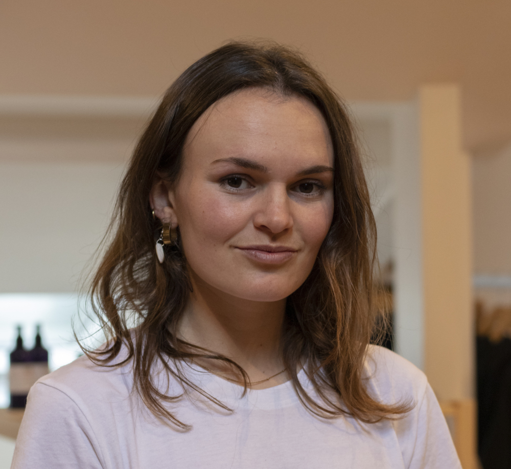 A woman with long brown hair is smiling slightly at the camera. She is wearing a white shirt and earrings, standing indoors with blurred shelves and bottles in the background.