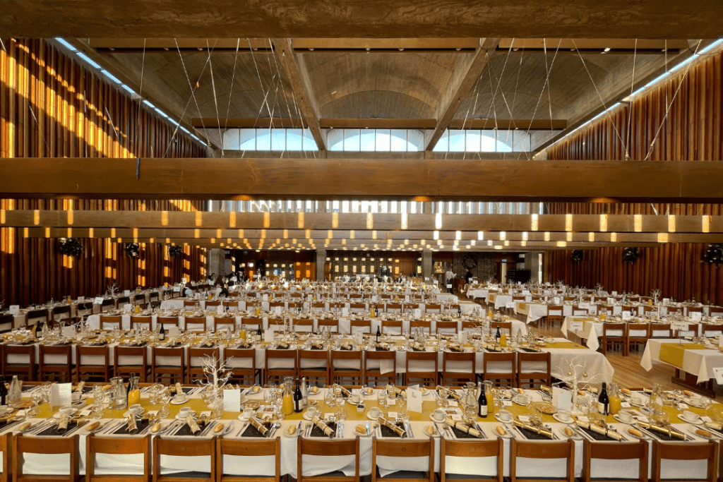 A large banquet hall filled with long tables set for a formal event. Tables are covered with yellow tablecloths and neatly arranged with plates, glasses, and cutlery. Warm sunlight streams in from large windows, illuminating the space.