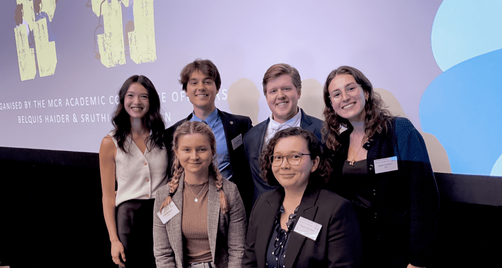 A group of six people, four women and two men, smiling and posing in front of a projection screen. They are dressed in business or smart casual attire, with conference name tags visible.