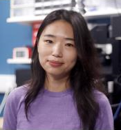 A woman with long dark hair is smiling slightly while standing in a room with a blue wall and shelves with electronic equipment. She is wearing a purple top.