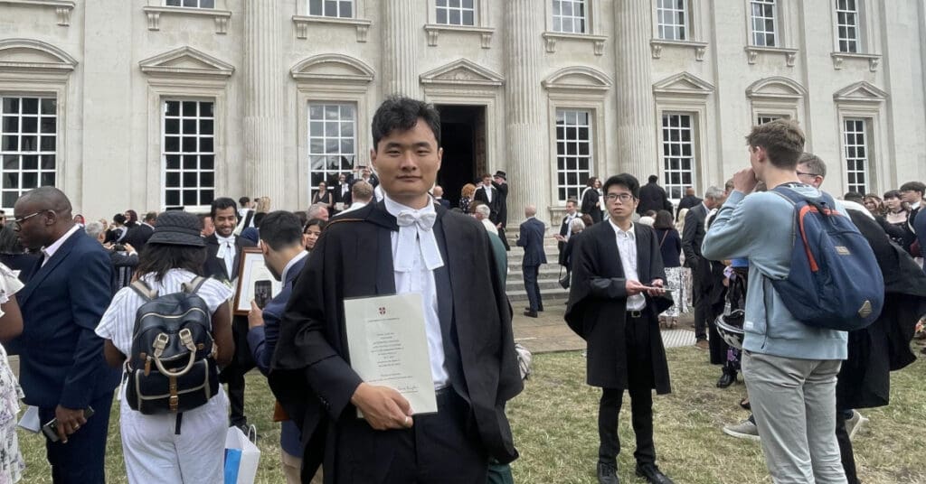 A graduate in academic attire holds a diploma in front of a historic building. People gather around, some in formal wear and others in casual clothes, celebrating and taking photos.