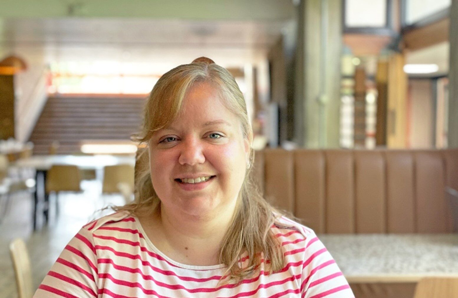 A woman with long hair smiles while seated in a restaurant with a striped top. The background shows a softly lit interior with wooden elements and empty chairs and tables, creating a cozy atmosphere.