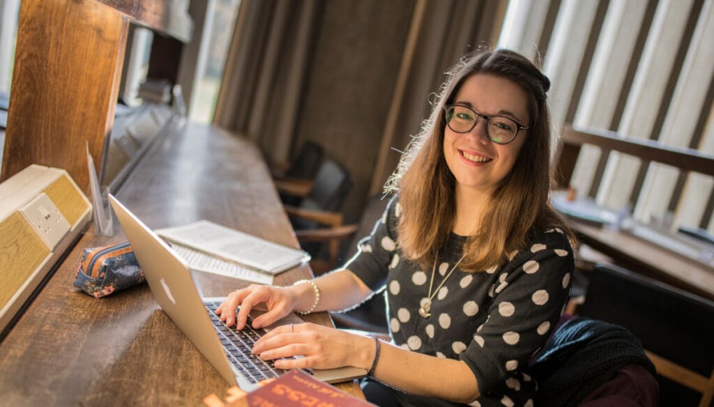 A woman with glasses and brown hair is smiling while typing on a laptop. She wears a black polka dot sweater and sits at a wooden desk with a notebook beside her. Soft light filters through nearby windows.