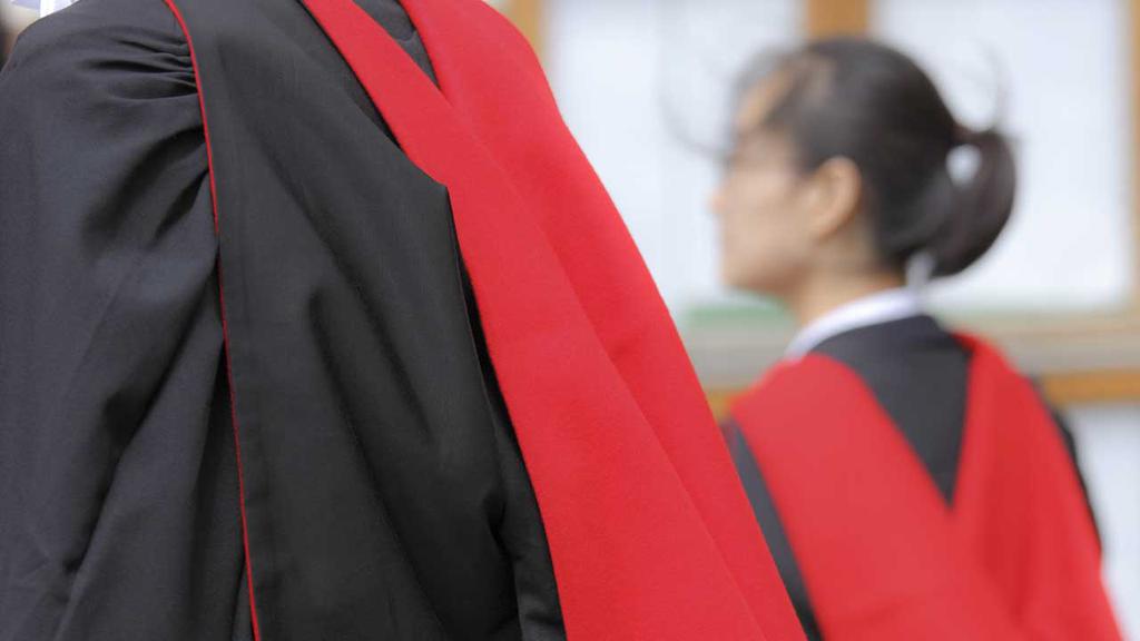 Two people are seen wearing black and red academic robes. The focus is on the robe of the person in the foreground, with the second person in the background slightly out of focus. The setting appears to be academic or ceremonial.