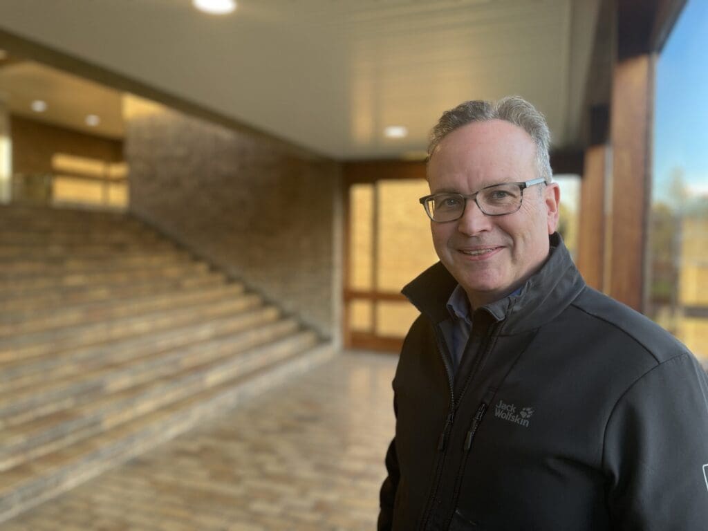 A man in a black jacket stands indoors beside a wooden staircase. He is smiling at the camera. The interior is warmly lit, featuring brick walls and a wooden ceiling.