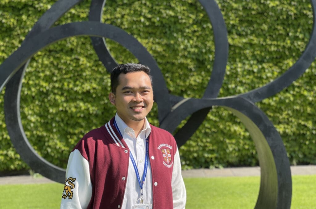 A person in a maroon and white jacket stands smiling in front of large metallic rings on a sunny day. The background is a lush green hedge.