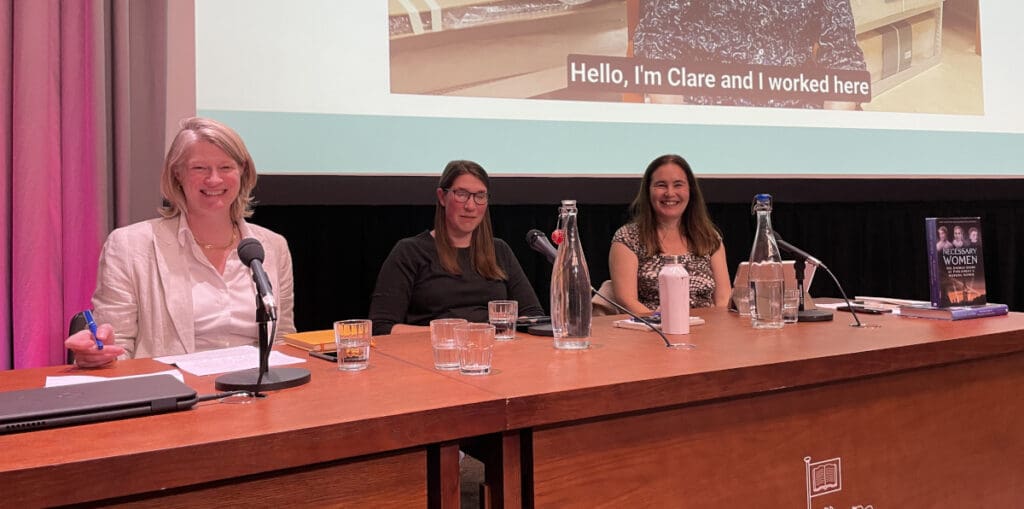 Three people sit at a panel table with microphones and water bottles. They are in a conference setting, smiling, with a presentation screen behind them.