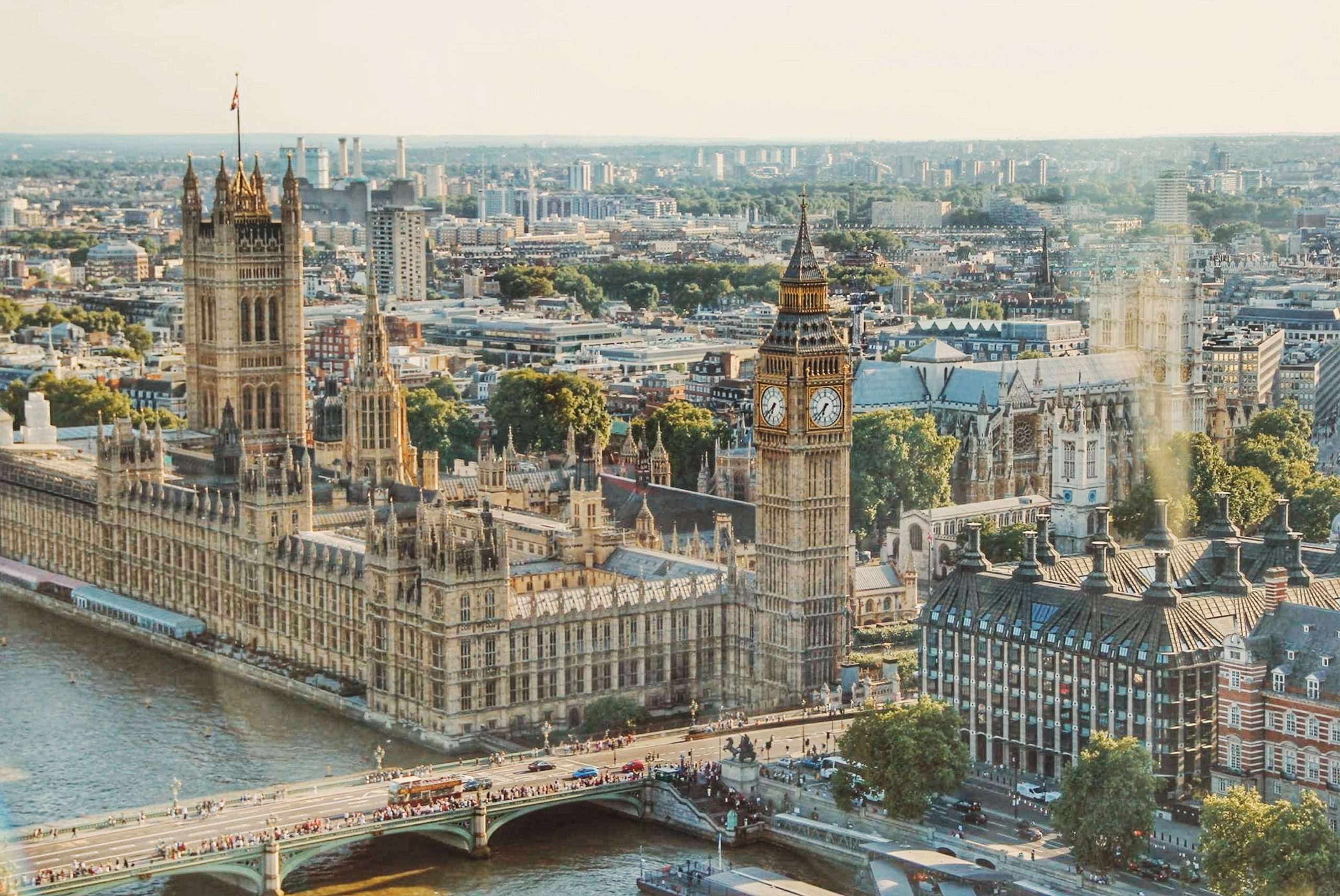 Aerial view of the Houses of Parliament and Big Ben in London, with the River Thames flowing alongside. Traffic and people are seen on the bridge, and the cityscape extends into the background under a clear sky.