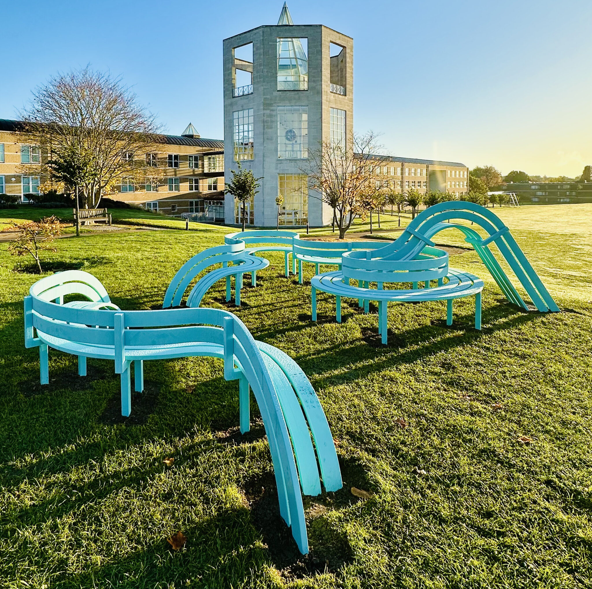 Modified Social Bench for Cambridge (2024), by Danish artist Jeppe Hein, a light blue bench which wraps around itself and into the grass