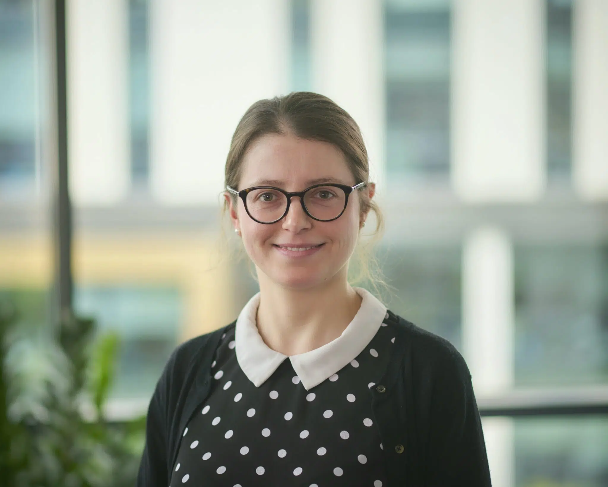 A person with glasses and long hair tied back is smiling indoors, wearing a polka-dot shirt and a cardigan. Large windows and a blurred background with greenery and buildings can be seen behind.