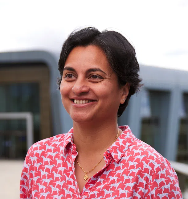 A person with short dark hair smiles while looking away from the camera, wearing a red shirt with white patterns. A modern building with a curved design is blurred in the background.